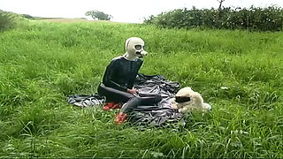 Fetish slave in latex walks and sucks a dildo on the grass